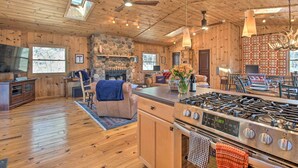 Une cabane spacieuse dotée d’une cheminée en pierre, de planchers en bois, d’une cuisine avec une cuisinière à gaz et d’un coin repas.