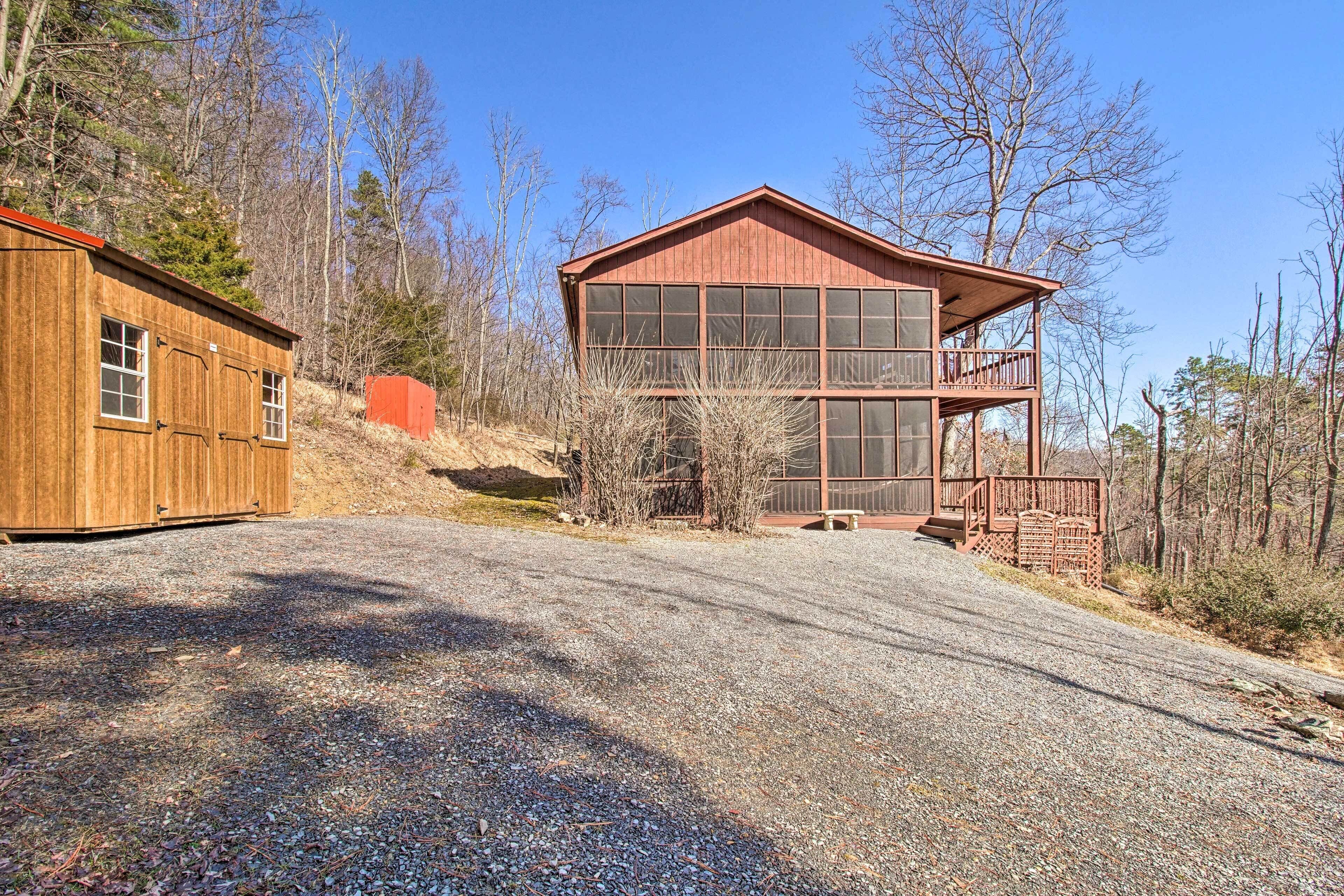 A two-story wooden cabin with large windows and a balcony, surrounded by trees and a gravel driveway.