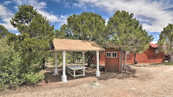 Une cabane rustique avec une table et un banc en bois, située sous un espace abrité et entourée d’arbres.