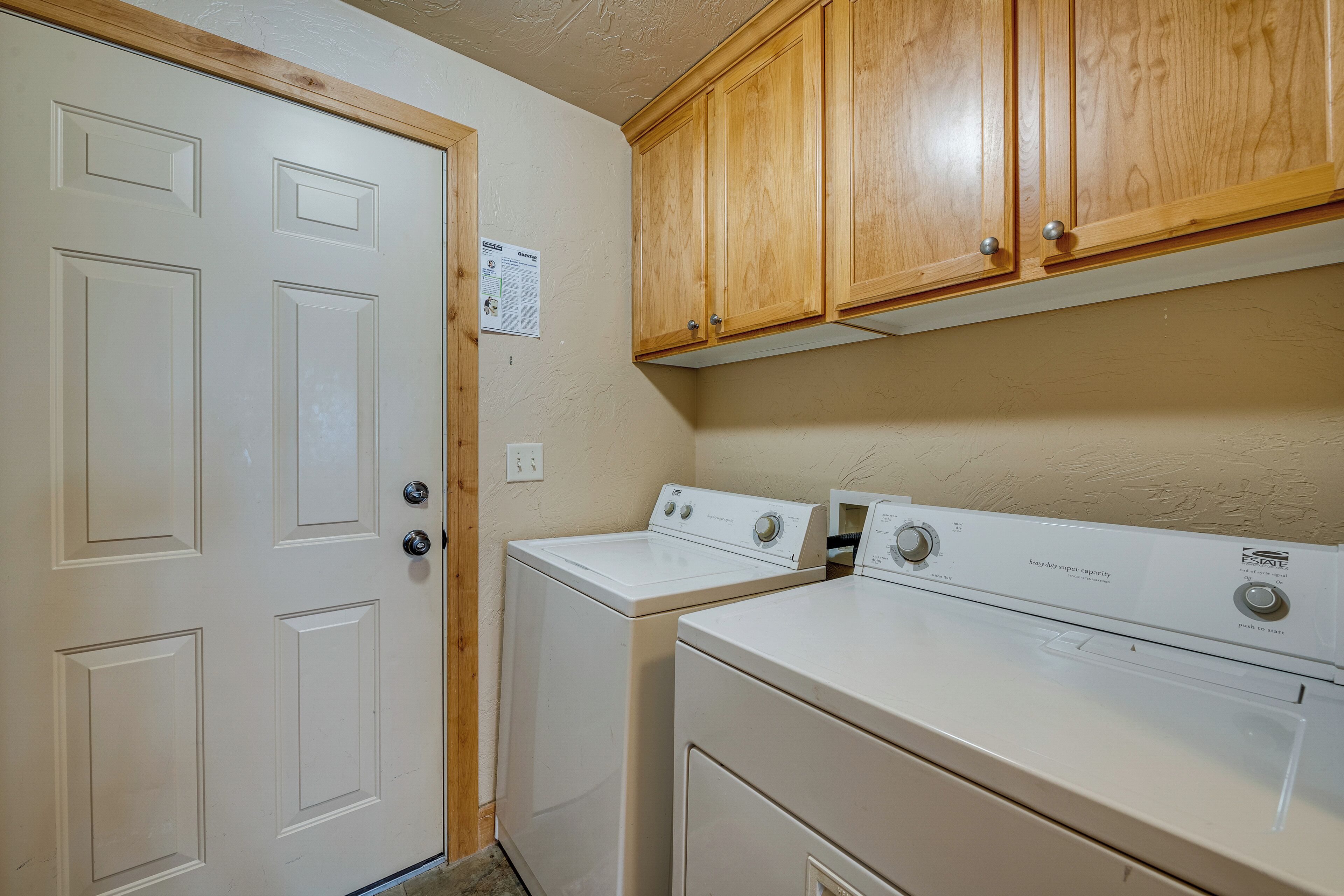 A laundry room with two white front-loading washers and dryers, wooden cabinets above, and a white door with a bulletin board.