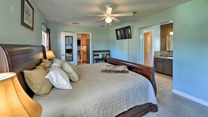 A bedroom with a large bed, a ceiling fan, a TV mounted on the wall, and a view into the adjacent kitchen.