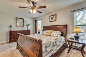 Une chambre à coucher avec un lit en bois, un ventilateur de plafond, une commode, une table de chevet, une lampe et une fenêtre avec des stores.