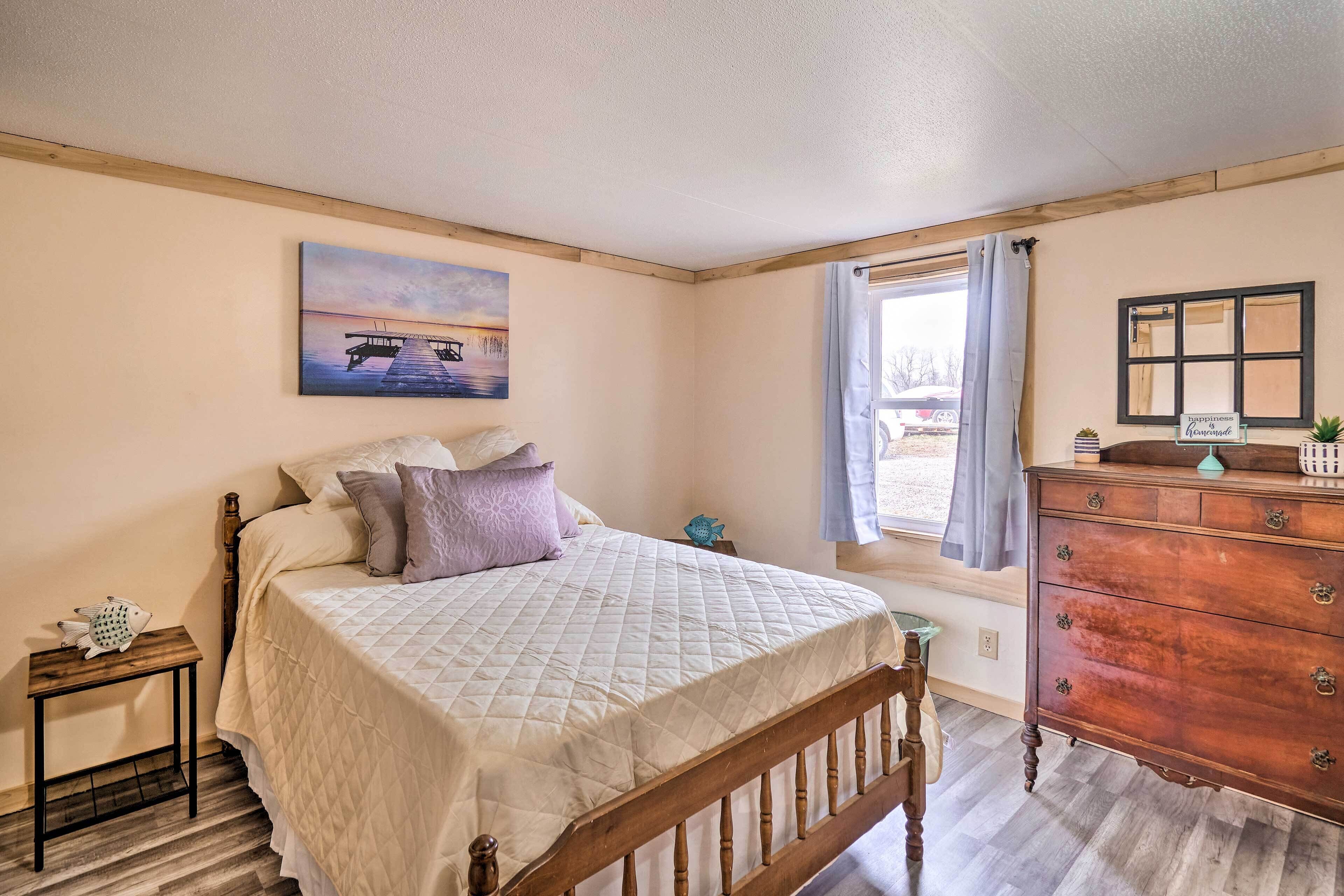 Een slaapkamer met een houten bed, nachtkastje, dressoir, een raam met gordijnen, en een schilderij aan de muur.