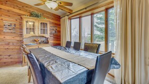 Interior de una cabaña de madera con una mesa de comedor, sillas, un ventilador de techo y una ventana con cortinas.
