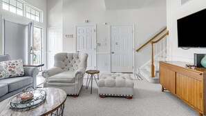 Een ruime woonkamer met een grijze bank, een gestoffeerde fauteuil, een houten salontafel en een witte voetenbankje. Er is een houten kast met een televisie erboven, en een trap met een houten leuning.