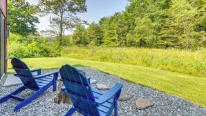 Zwei blaue Adirondack-Stühle auf einer Schotterterrasse mit einem gepflegten Rasen und dichtem Wald im Hintergrund.