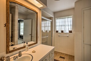 A bathroom with a large mirror, a sink, a shower, and a window with curtains.