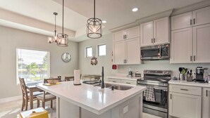 A modern kitchen with white cabinetry, a central island, stainless steel appliances, and a dining area with chairs and a table. - Stunning Laguna Vista Retreat w/ Pool & Grill (Port Isabel)