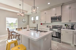 A modern kitchen with white cabinetry, a central island, stainless steel appliances, and a dining area with chairs and a table. - Stunning Laguna Vista Retreat w/ Pool & Grill (Port Isabel)