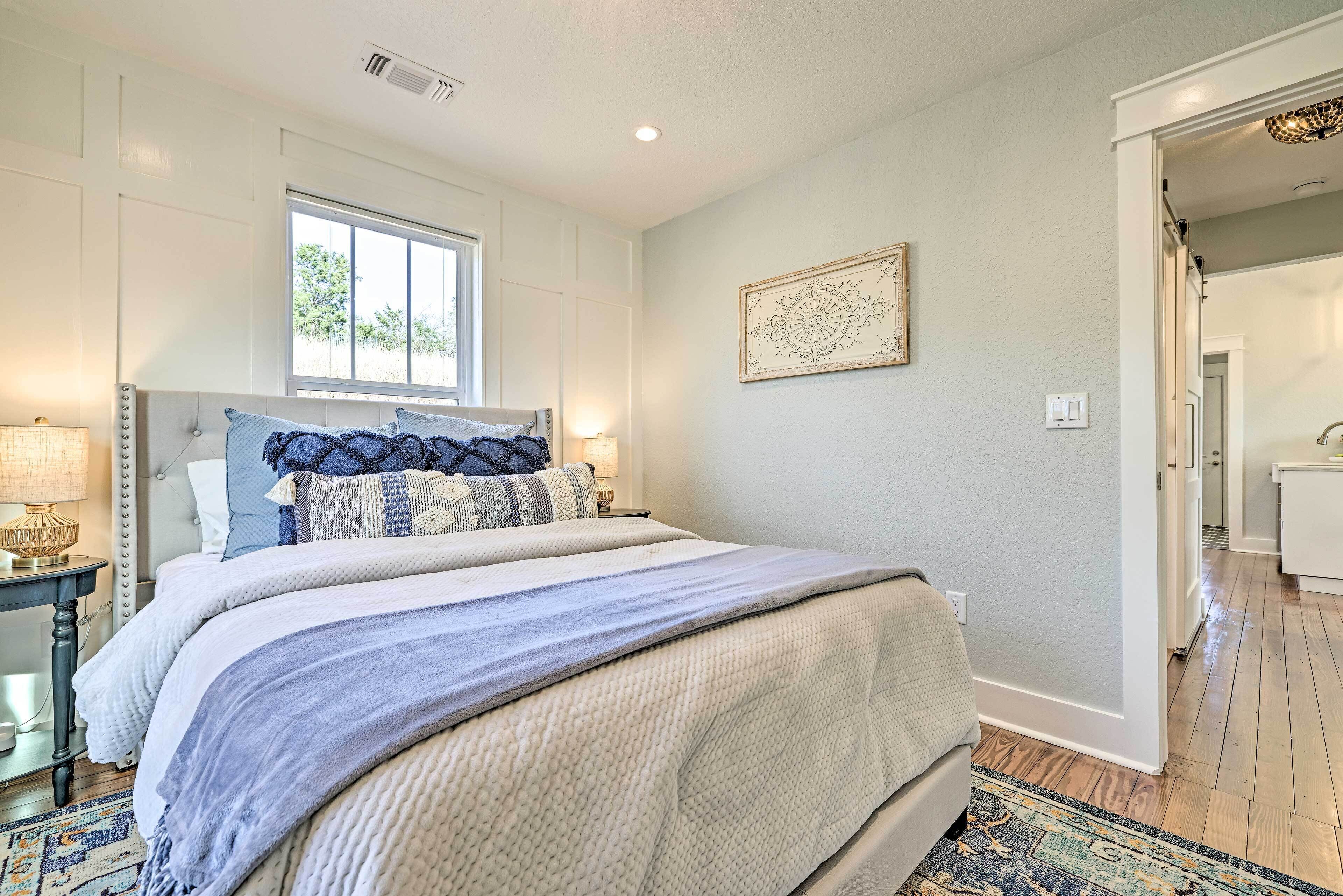 A bedroom with a large bed, a nightstand, a window, and a door leading to another room.