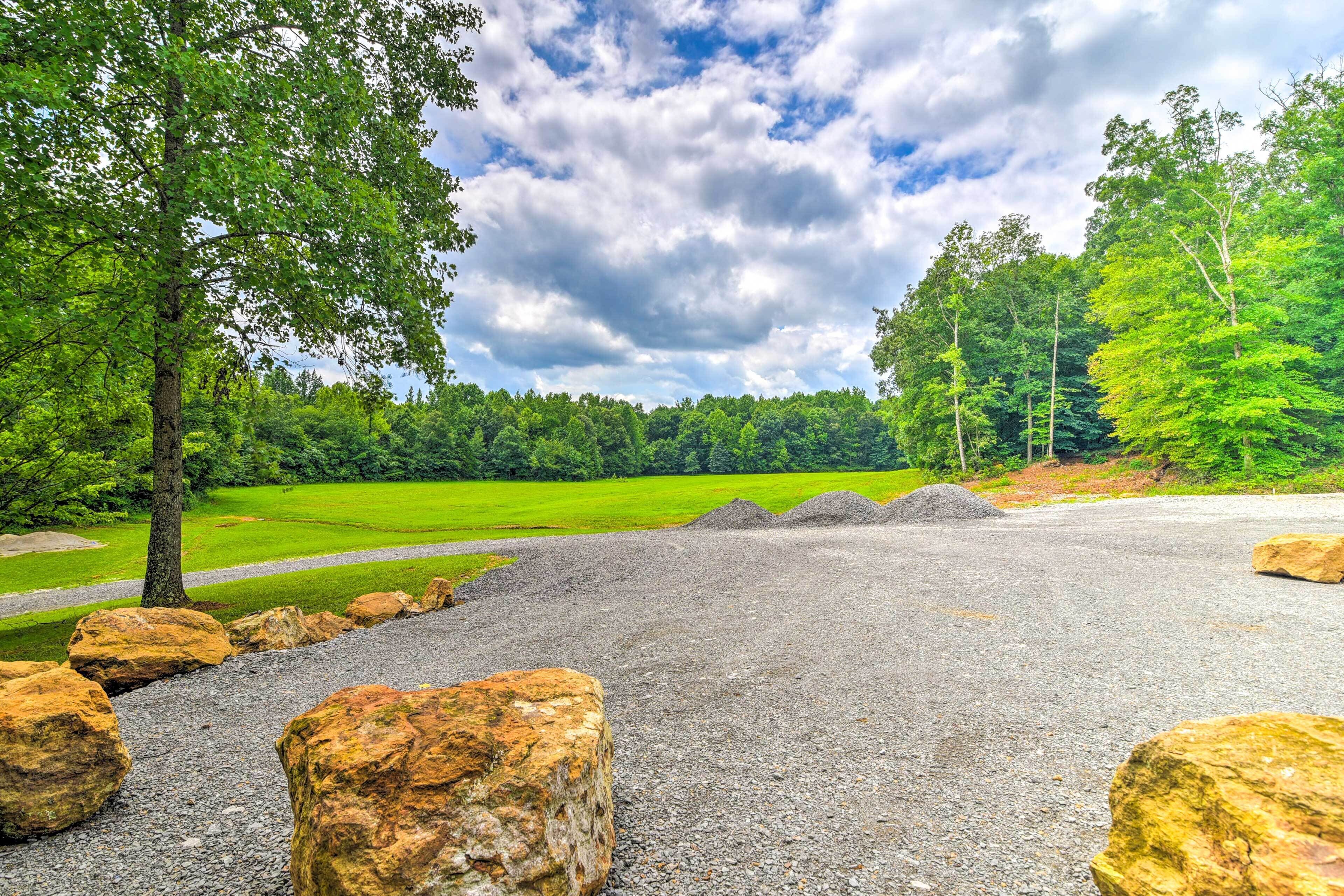 A gravel area with large rocks, a grassy field, and trees under a partly cloudy sky.