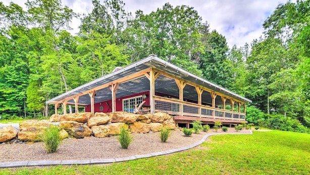 A lodge with a covered deck and a stone foundation, surrounded by a well-maintained lawn and dense forest. - Secluded Brownsville Cabin w/ Deck & Fire Pit (Roundhill)
