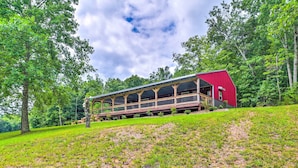 A red-roofed building with a covered deck, surrounded by trees and grassy terrain. - Secluded Brownsville Cabin w/ Deck & Fire Pit (Roundhill)