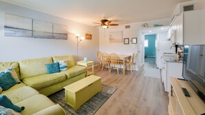 Un salon moderne avec un canapé vert, une table à manger avec des chaises, un coin cuisine équipé d’un réfrigérateur et un ventilateur de plafond.