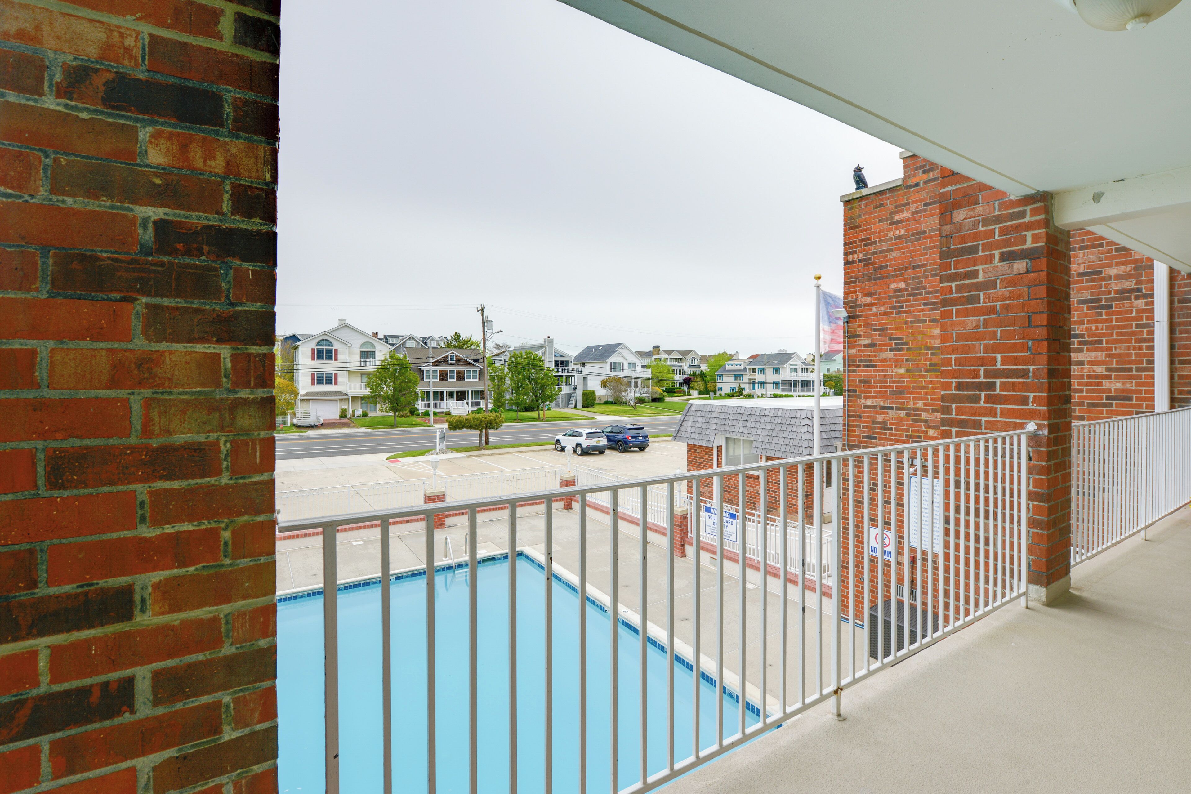 A view from a balcony with a swimming pool, brick walls, and a parking area with cars.