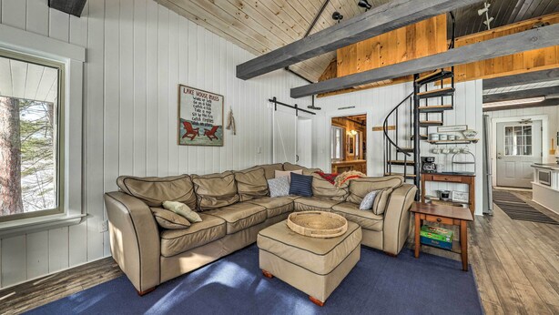 Ein großzügiges Wohnzimmer mit einem großen, beigefarbenen Ecksofa, einem kleinen Hocker und einem Holztisch. Eine Wendeltreppe führt auf eine obere Ebene, und im Hintergrund ist eine Küchenzeile zu sehen.