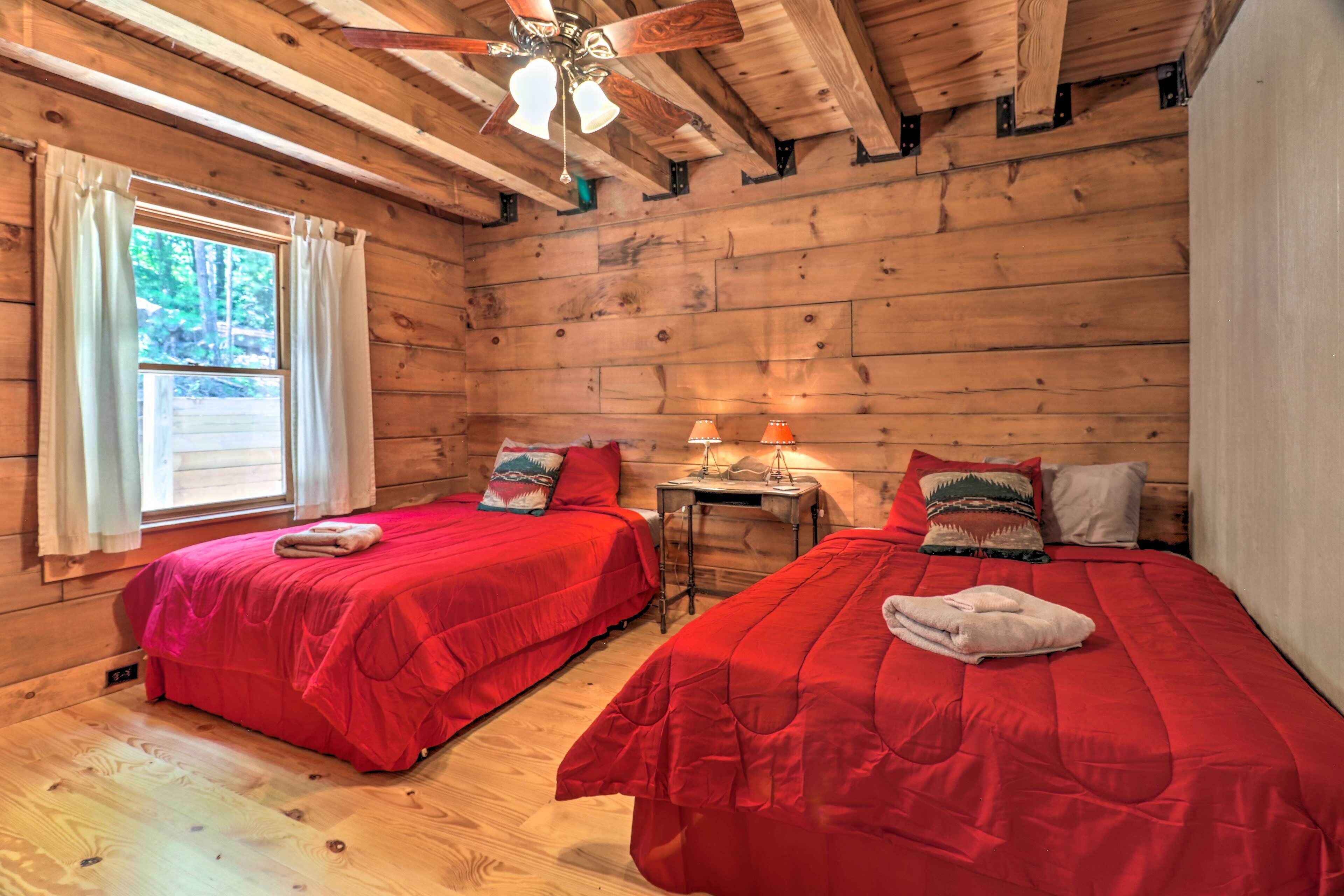 A cabin with two beds, wooden walls, a ceiling fan, a window with curtains, and a small table with a lamp.
