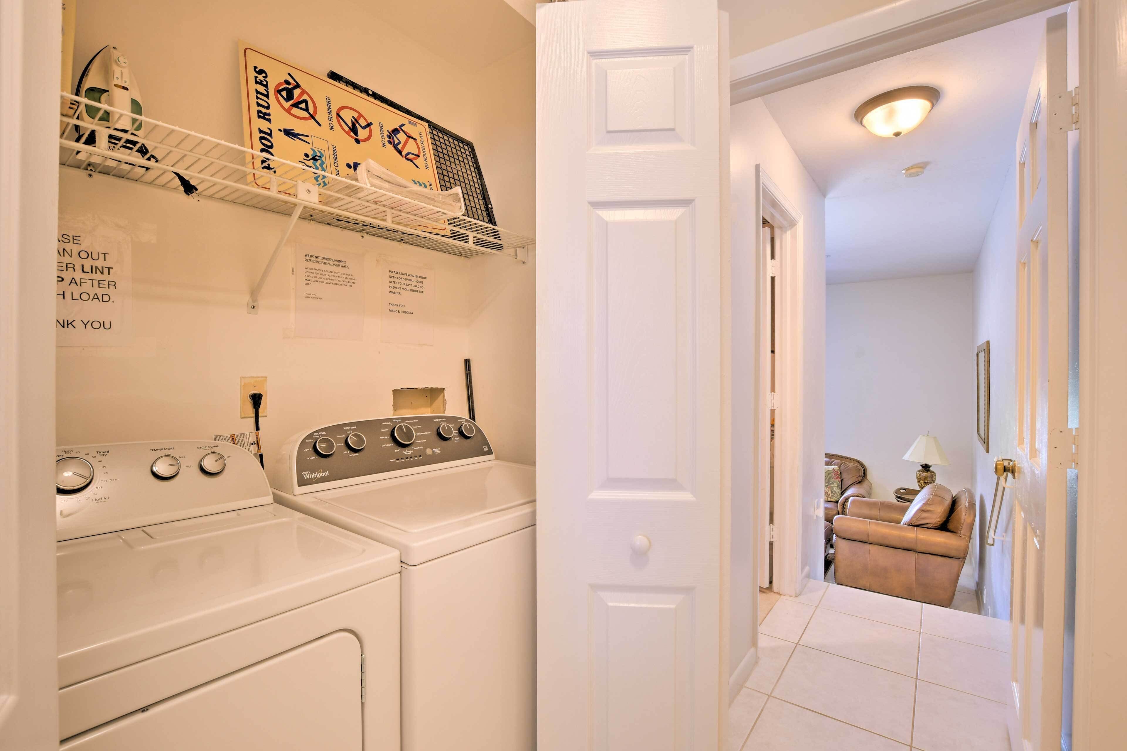 A laundry room with a Whirlpool washer and dryer, a shelf with laundry supplies, and a door leading to a living area with a couch and a lamp.