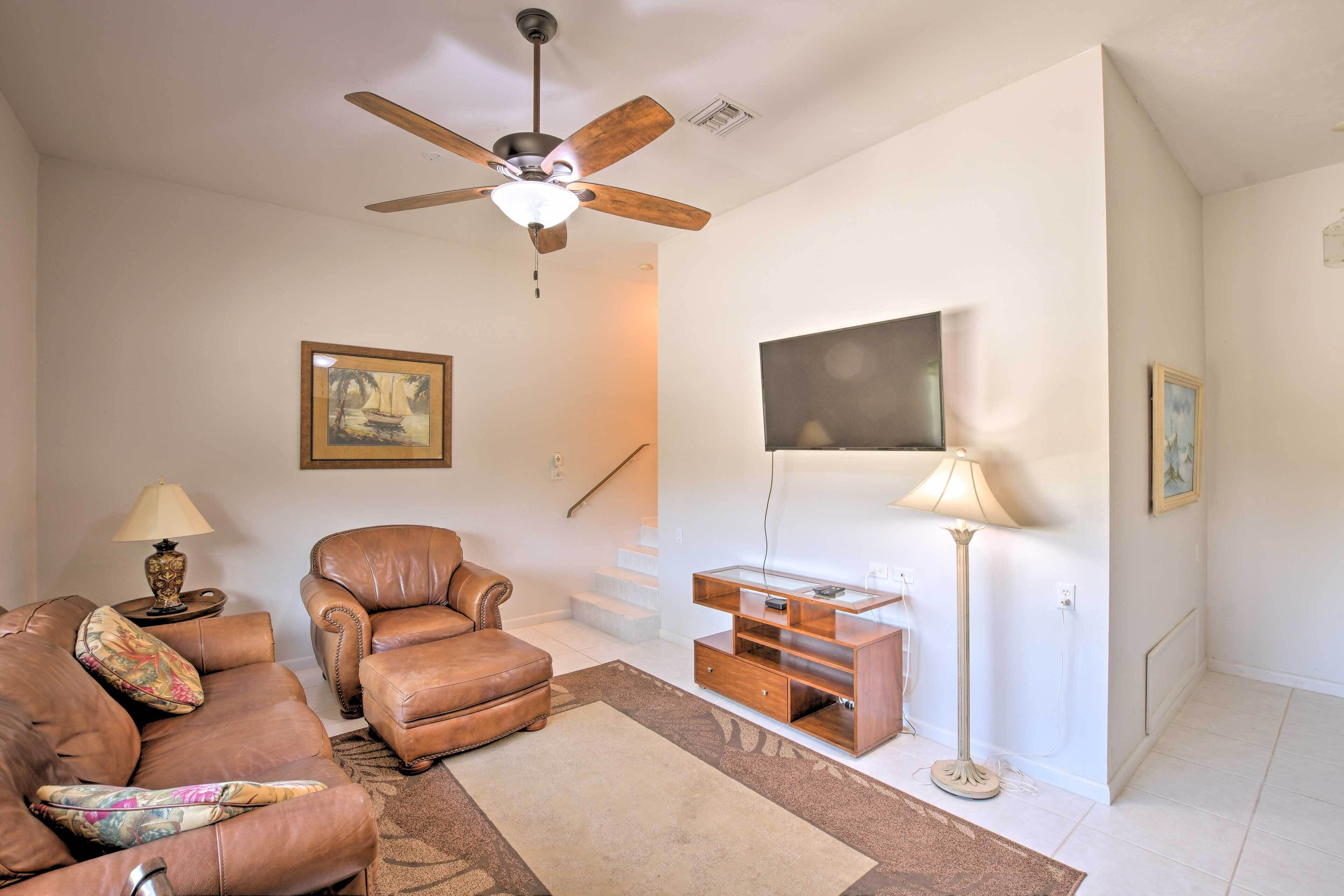A living room with a ceiling fan, leather sofa, a glass coffee table, and a flat-screen TV mounted on the wall.