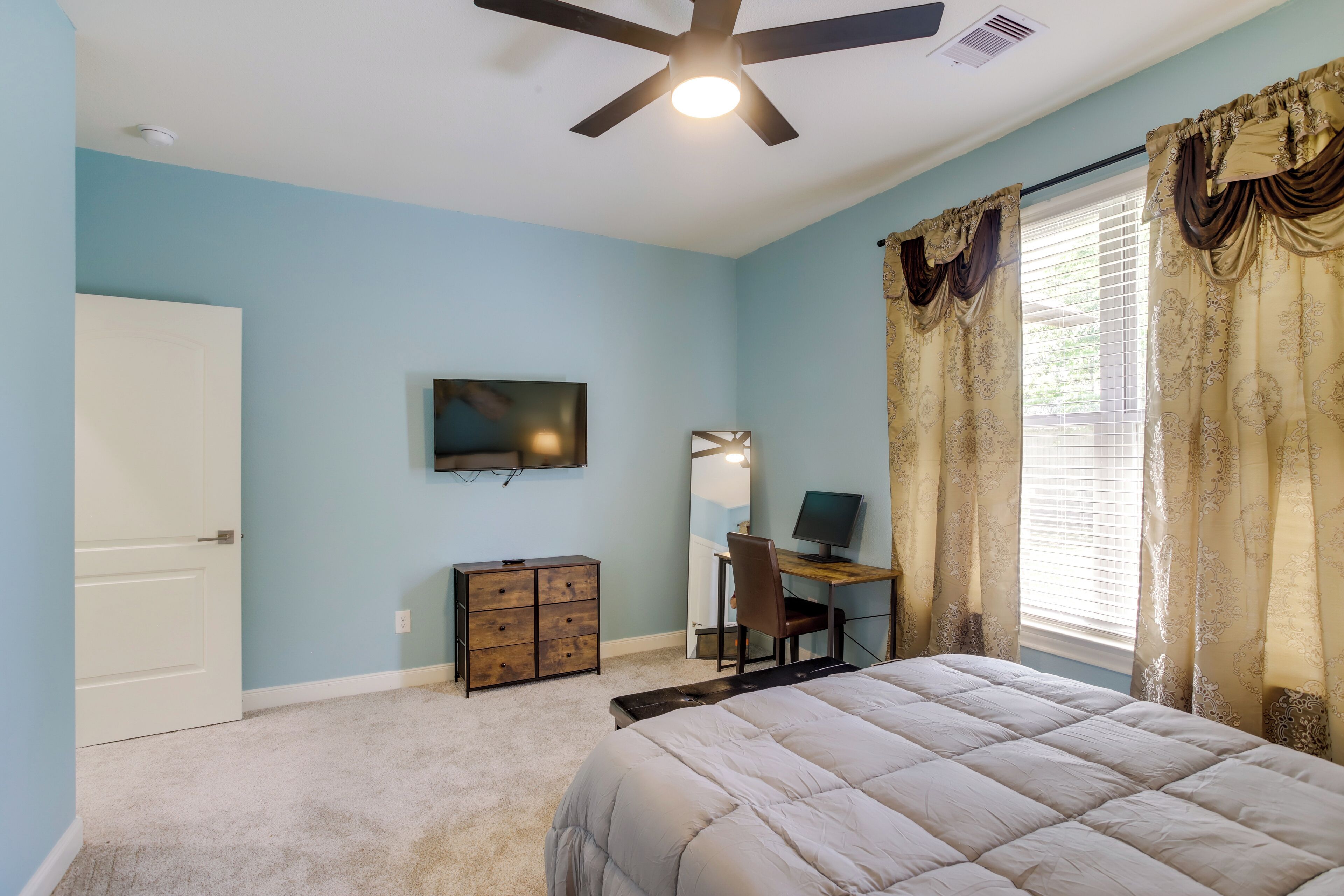 A bedroom with a bed, a desk with a computer, a chair, a dresser, a television, and a ceiling fan.