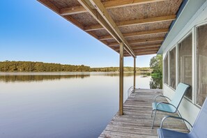 A wooden deck with a covered area and two chairs, overlooking a calm lake with a forested shoreline. - Lakefront Crockett Cabin w/ Boathouse & Kayaks! (Grapeland)