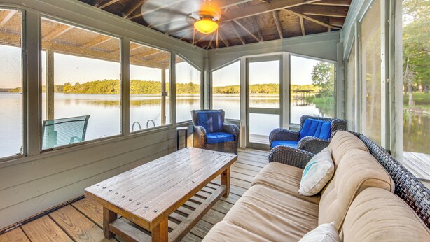 A covered outdoor seating area with a wooden table and cushioned seating, overlooking a lake.