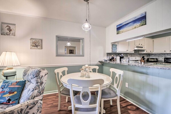 A dining area with a round table and chairs, a hanging light fixture, and a kitchen with white cabinets and a marble countertop.