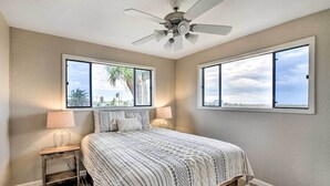 Un dormitorio con cama, dos lámparas de noche, ventilador de techo y ventanas amplias con vista a palmeras y al océano.
