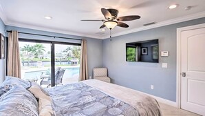 Une chambre avec un lit, un ventilateur de plafond, une grande fenêtre avec des rideaux, une chaise et une vue sur une piscine et des palmiers.