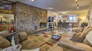 Una sala de estar acogedora con pared de piedra, sofá de cuero y una mesa de centro de vidrio. Se ve una cocina con taburetes de bar y un televisor al fondo.