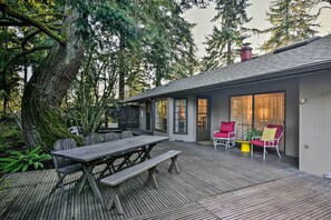 A wooden deck with a picnic table and chairs, surrounded by trees and a building with large windows and a door. - Spacious Hillsboro Home ~ 18 Mi to Portland! (Hillsboro)