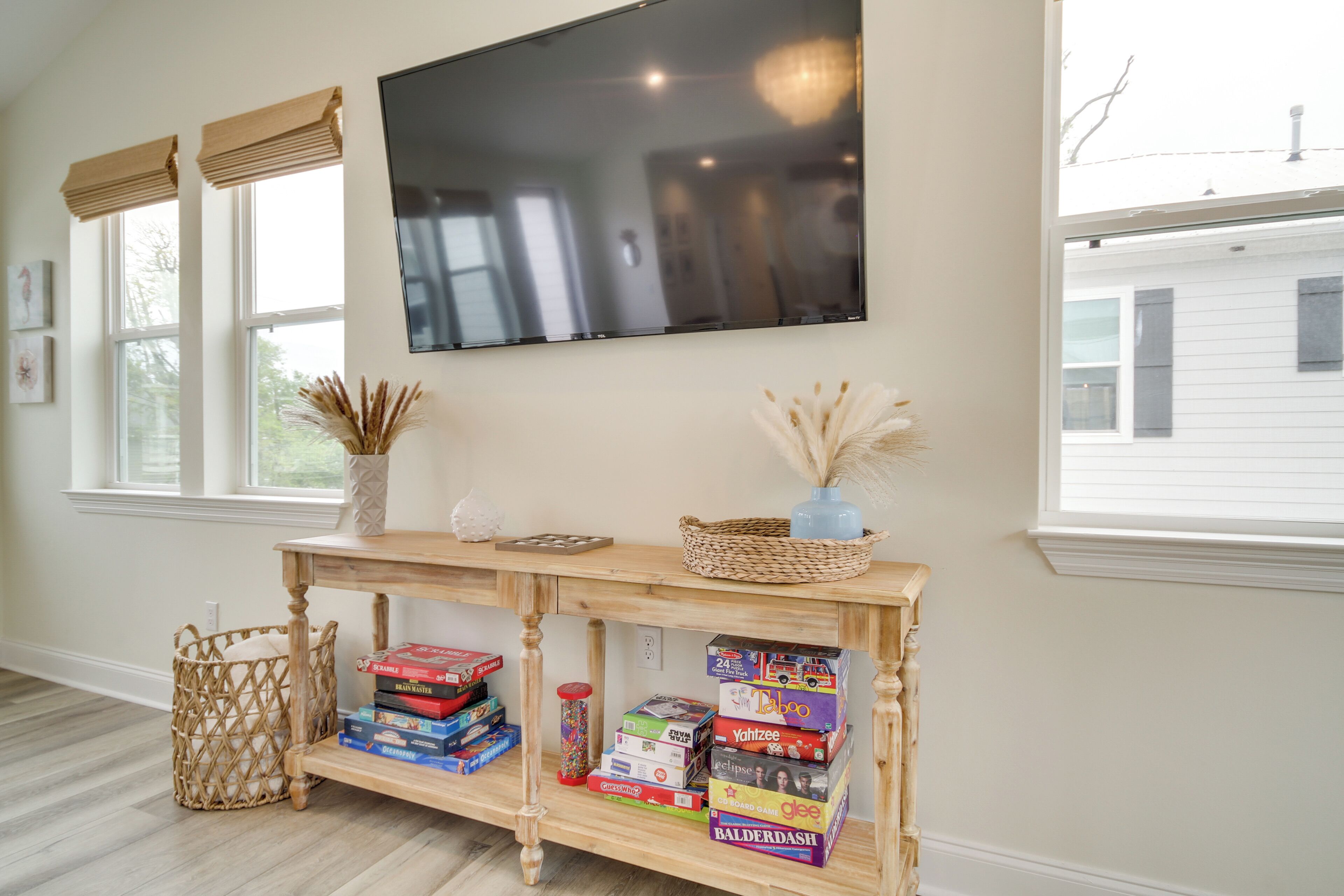 A living room with a wooden console table displaying board games, a flat-screen TV mounted above it, and a large window with blinds.