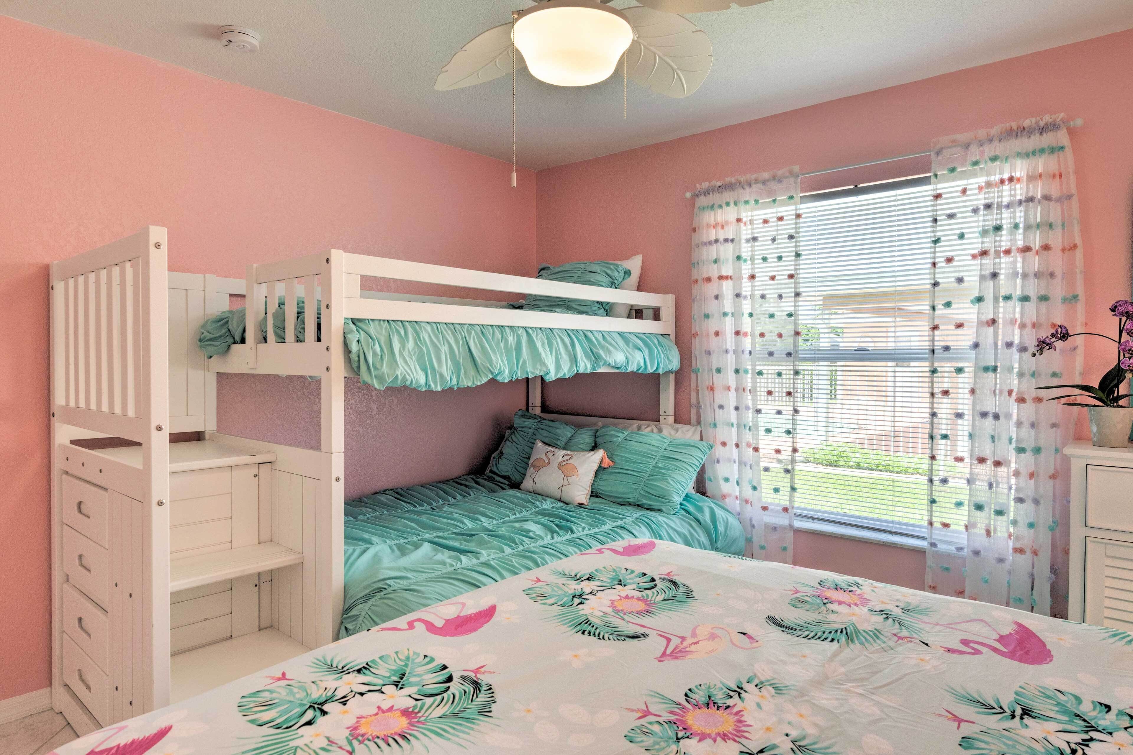 Un dormitorio con decoración rosa que incluye una cama nido, ventilador de techo y una ventana con cortinas coloridas.