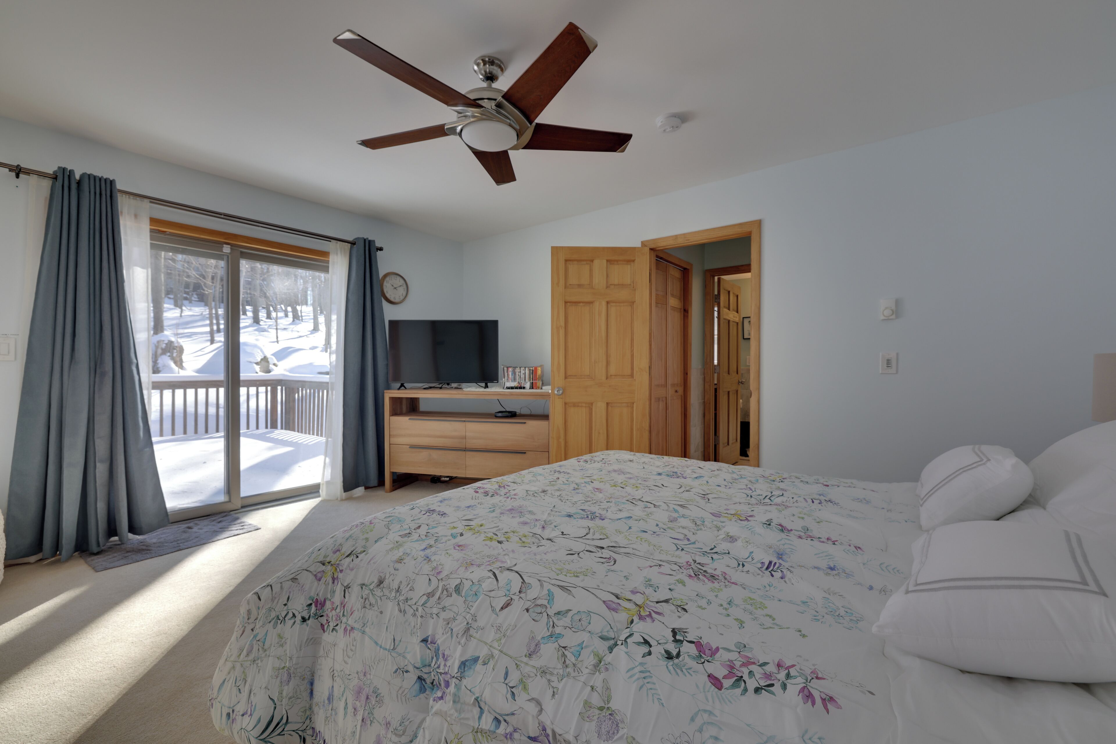 A bedroom with a bed, a ceiling fan, a TV, a wooden wardrobe, and a sliding glass door leading to a balcony.