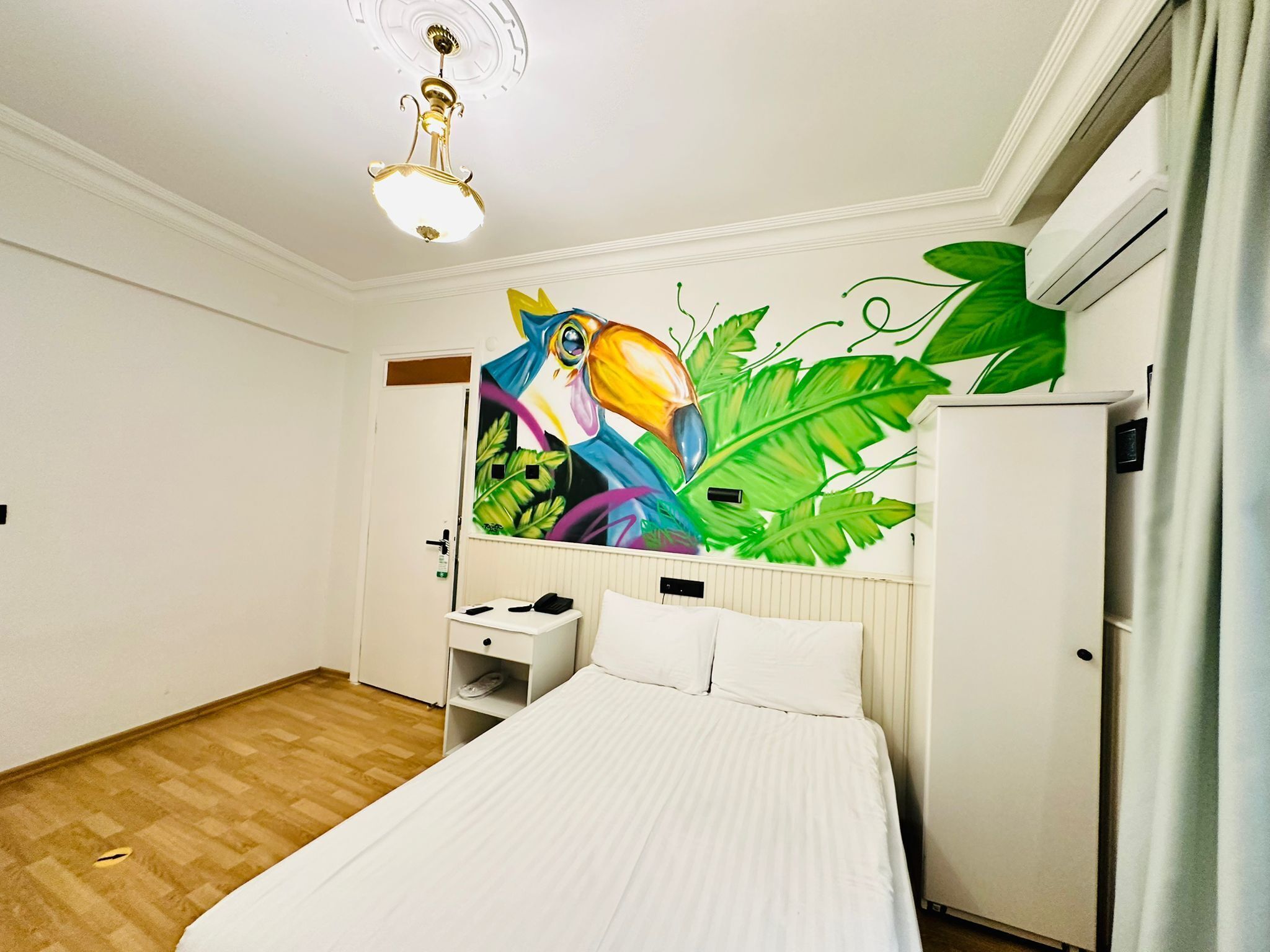 A bedroom with a bed, a desk, and a wall mural of a toucan among tropical leaves.