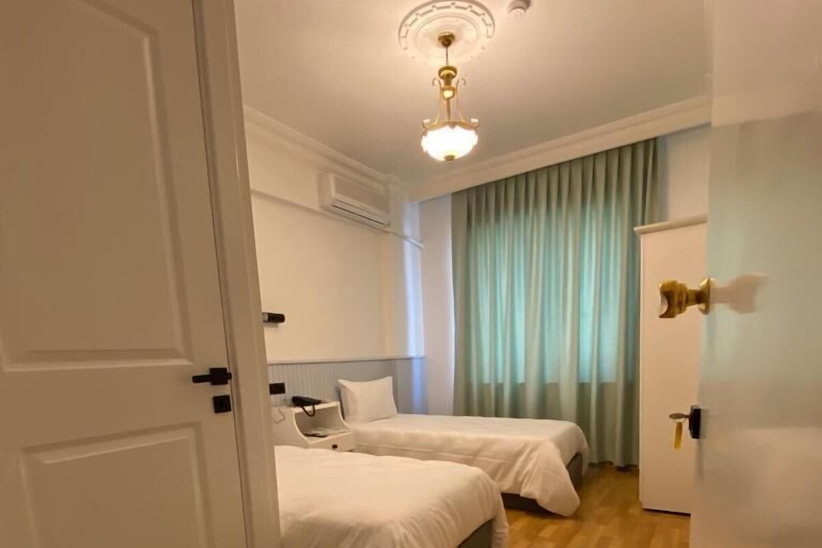 A hotel room with two beds, a ceiling light fixture, a window with curtains, and a door.