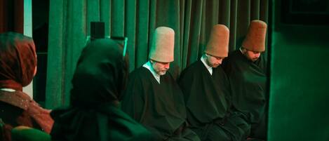 Un grupo de personas vestidas con atuendo tradicional sentadas en una habitación con cortinas verdes y paneles de madera.