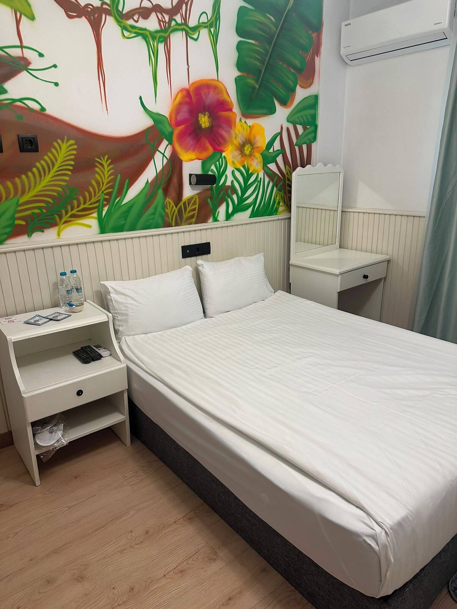 A hotel room with a bed, a bedside table with a phone and water bottles, a mirror, and a wall mural with tropical leaves and flowers.