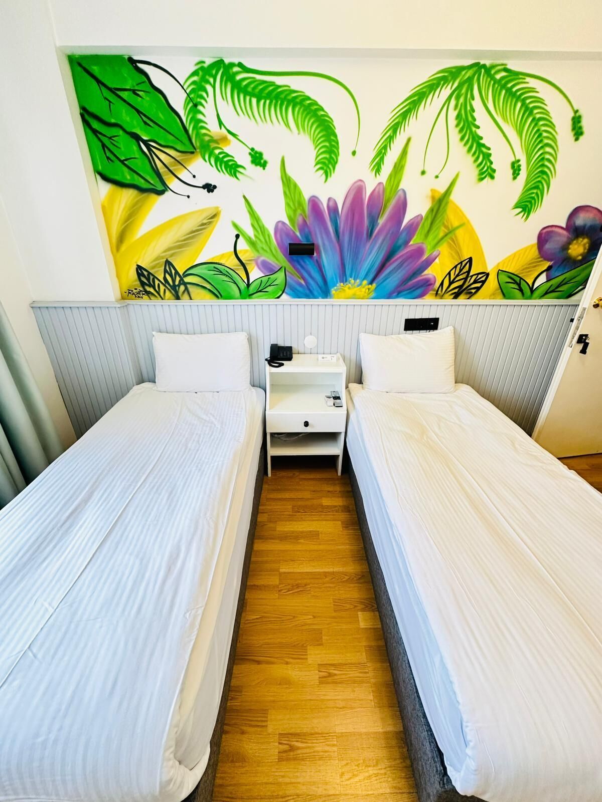 Quarto de hotel com duas camas de solteiro, uma pequena mesa de cabeceira com telefone e um mural colorido na parede.