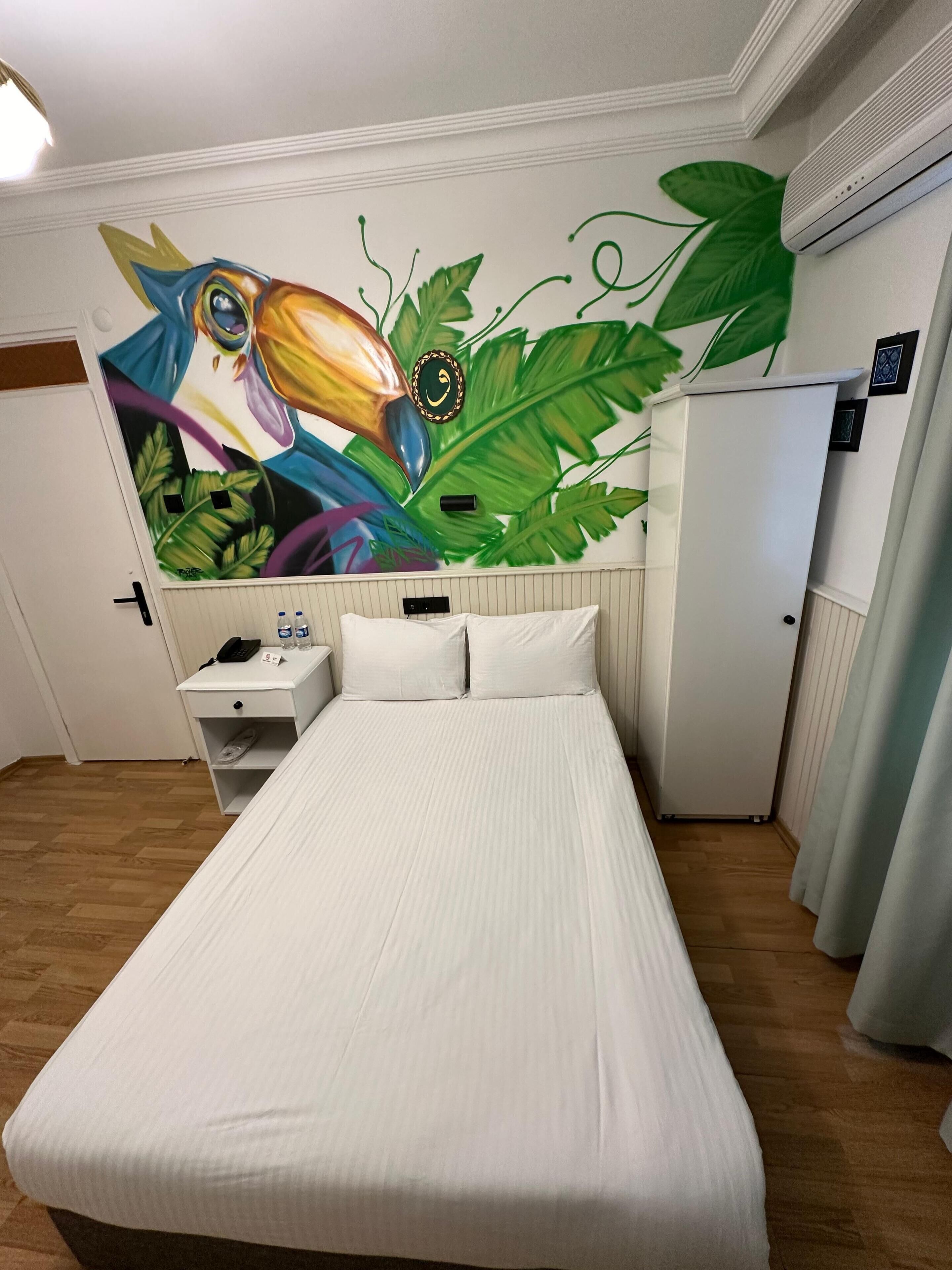 Une chambre d’hôtel avec un lit, une table de chevet, un bureau et une fresque murale représentant un toucan.