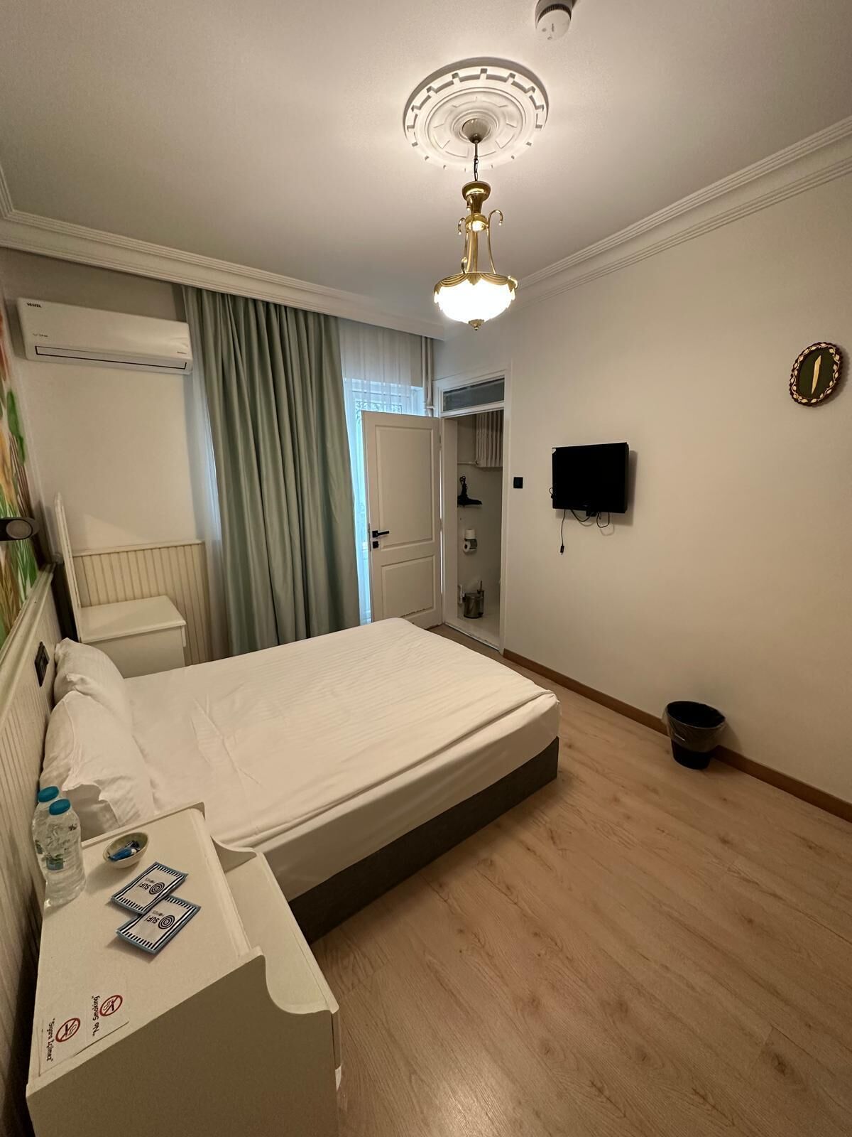 Een hotelkamer met een bed, een plafondlamp, een airconditioningapparaat, een aan de muur bevestigde televisie en een kleine prullenbak.