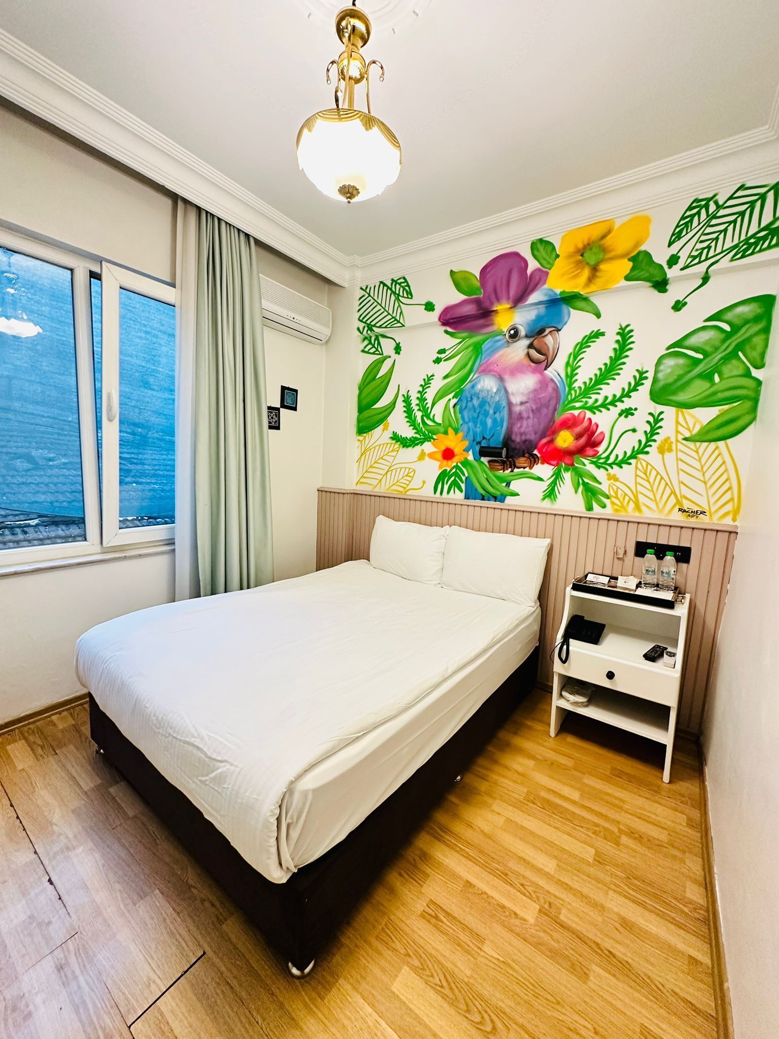 A bedroom with a bed, a nightstand, a wall mural, a window, and a ceiling light.
