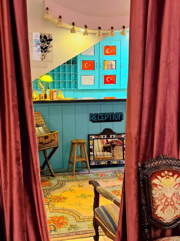 A reception area with a blue wall, a framed mirror, a wicker chair, a wooden stool, and a reception sign. - Sufi Homes (Konya)