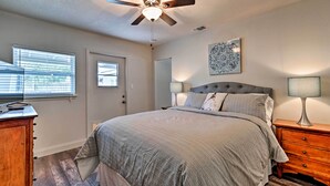 Un dormitorio con cama, ventilador de techo, mesita de noche, lámpara, cómoda, puerta y ventana con persianas.