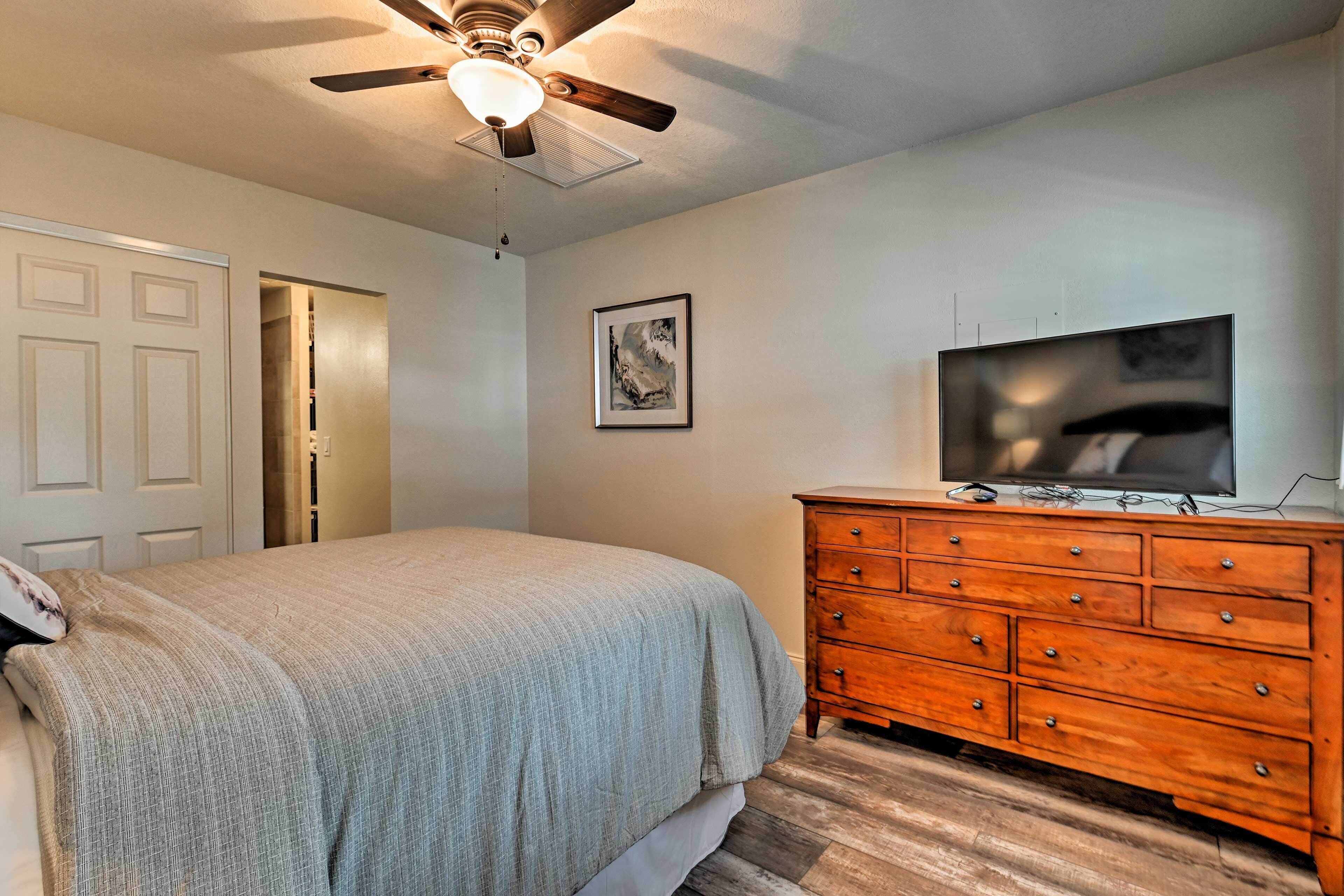 Une chambre à coucher comprenant un lit, une commode, un ventilateur de plafond, un téléviseur et une porte.