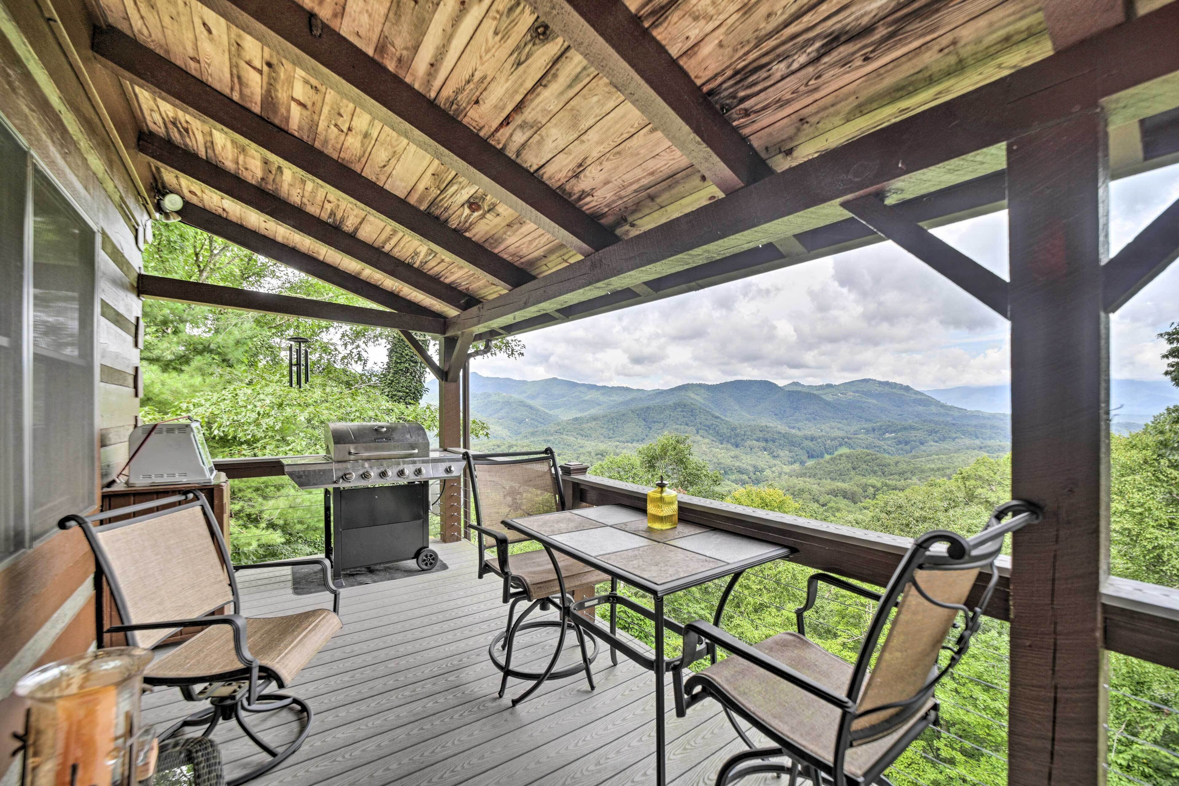 A covered outdoor deck with a table and chairs, a grill, and a view of mountains.