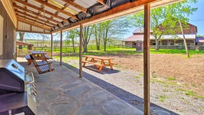 En udendørs overdækket terrasse med grill, picnicbord og udsigt til en gård og lade i baggrunden.