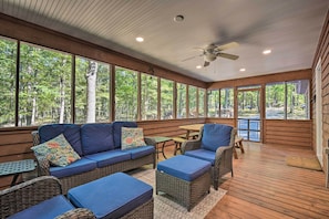 A covered outdoor seating area with wicker furniture, a ceiling fan, and large windows offering views of a forested area. - Cacapon River Cabin w/ Private River Access! (Great Cacapon)