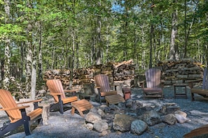 A forested area with Adirondack chairs, a fire pit, and a stack of firewood. - Cacapon River Cabin w/ Private River Access! (Great Cacapon)