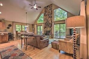 A spacious living room with a stone fireplace, large windows, and a ceiling fan. - Cacapon River Cabin w/ Private River Access! (Great Cacapon)